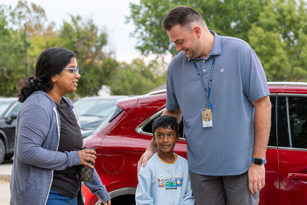 A Putnam City staff member smiles while greeting a parent and young student beside a red vehicle during morning drop-off.