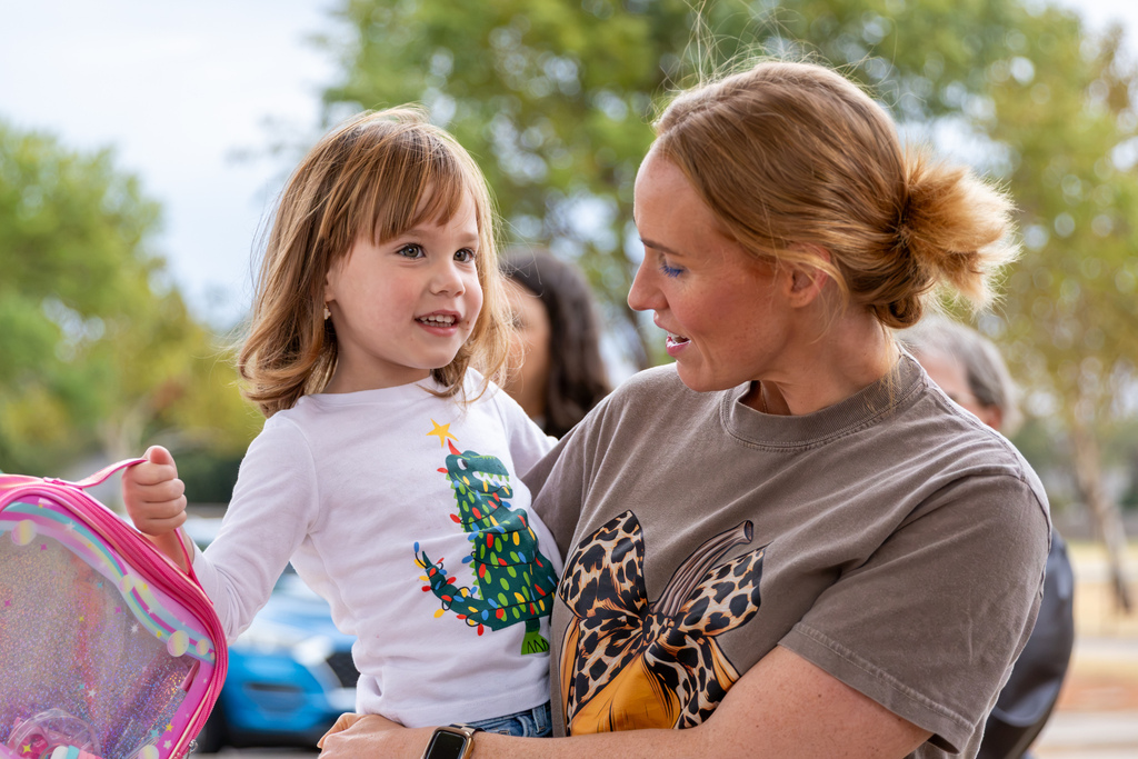 A mother holds her young daughter, who carries a colorful pink backpack, as they share a moment together outside the school.