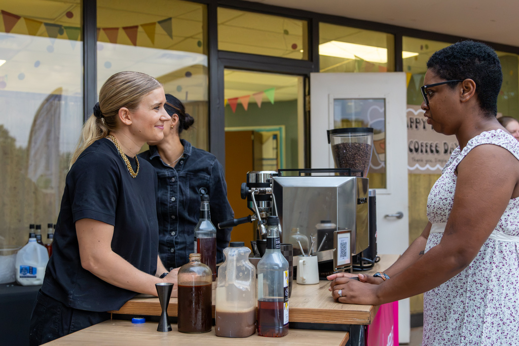 A coffee vendor smiles while speaking with a woman across an outdoor espresso station stocked with coffee syrups and supplies.