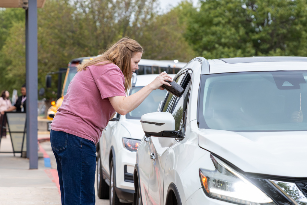 A woman hands a black Simple Modern tumbler through the window of a white SUV during morning drop-off.
