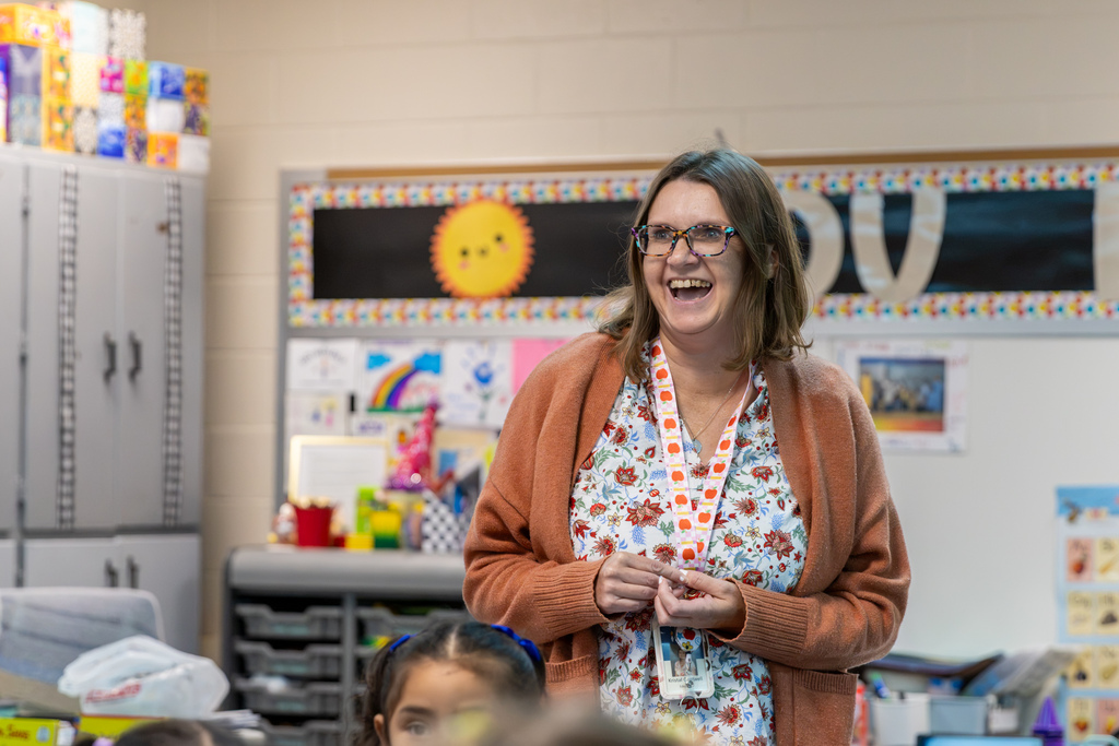 Lake Park Elementary teacher Kristal Cantwell laughs in her classroom following her surprise Teacher of the Month recognition. She wears a rust-colored cardigan and stands in front of a bulletin board decorated with student artwork.