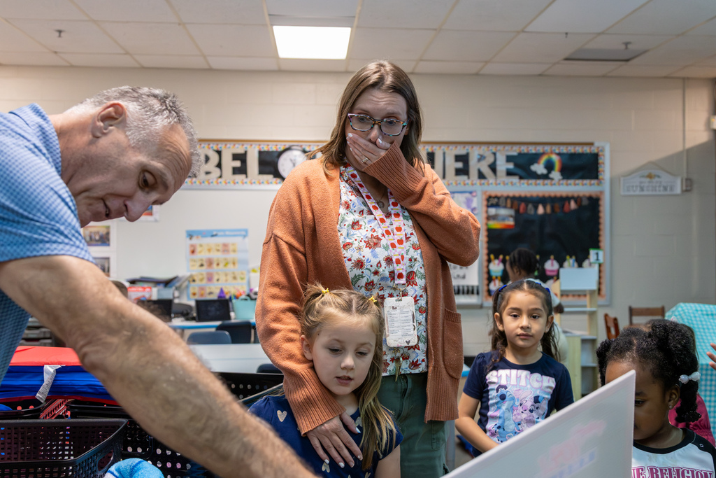 Kristal Cantwell stands in her classroom with her hand over her mouth as a visitor prepares to present her Teacher of the Month recognition. Several students watch the surprise unfold.