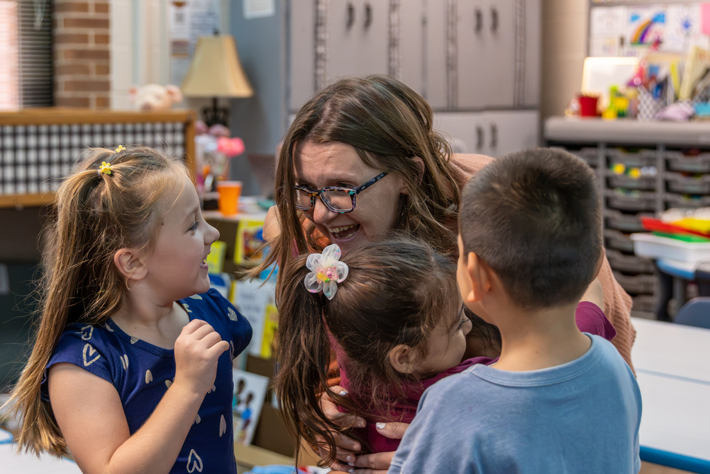 Kristal Cantwell smiles and leans toward three excited students in her classroom. One student wraps her arms around Cantwell while another smiles at her during the surprise celebration.
