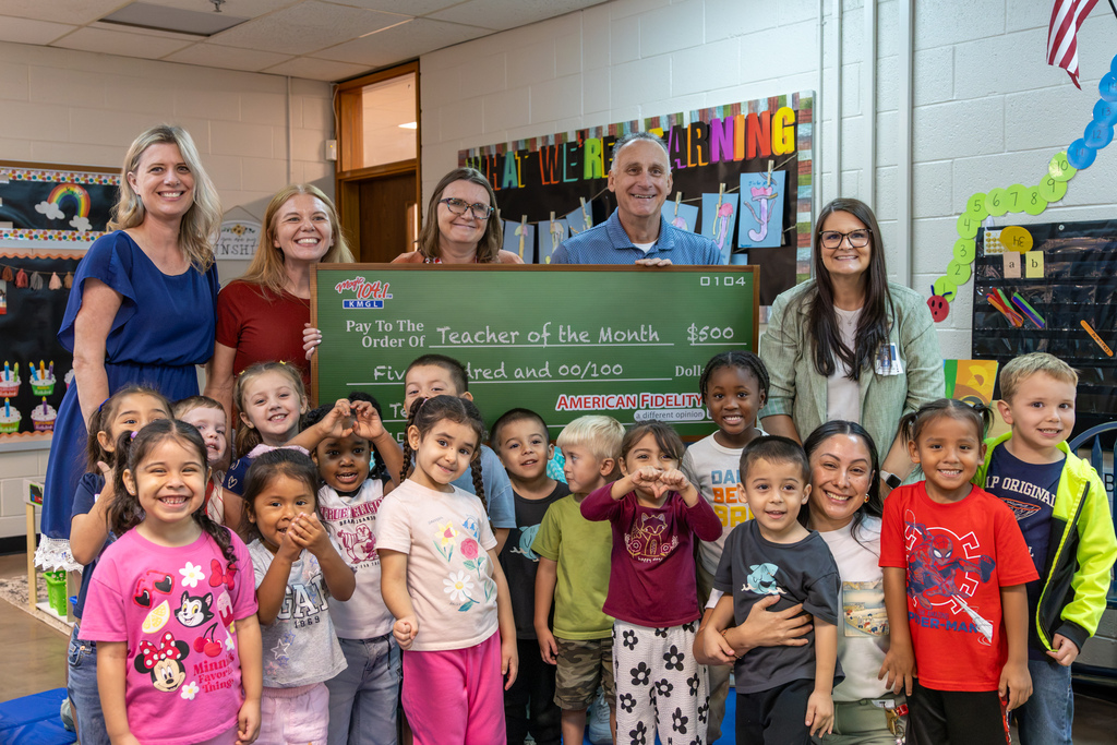 Kristal Cantwell poses with her students and four adults in her classroom behind an oversized Magic 104.1 Teacher of the Month check for $500. Students smile, wave, and make heart shapes with their hands.