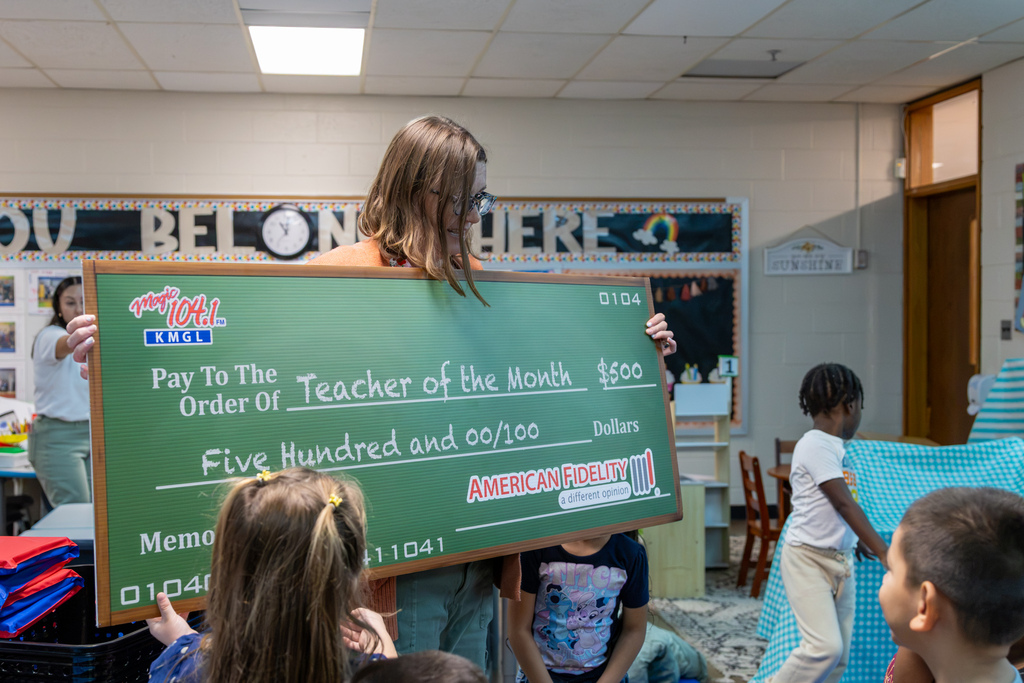 Kristal Cantwell holds an oversized green Magic 104.1 Teacher of the Month check for $500 in her classroom as students gather around her. American Fidelity is listed as a sponsor on the check.