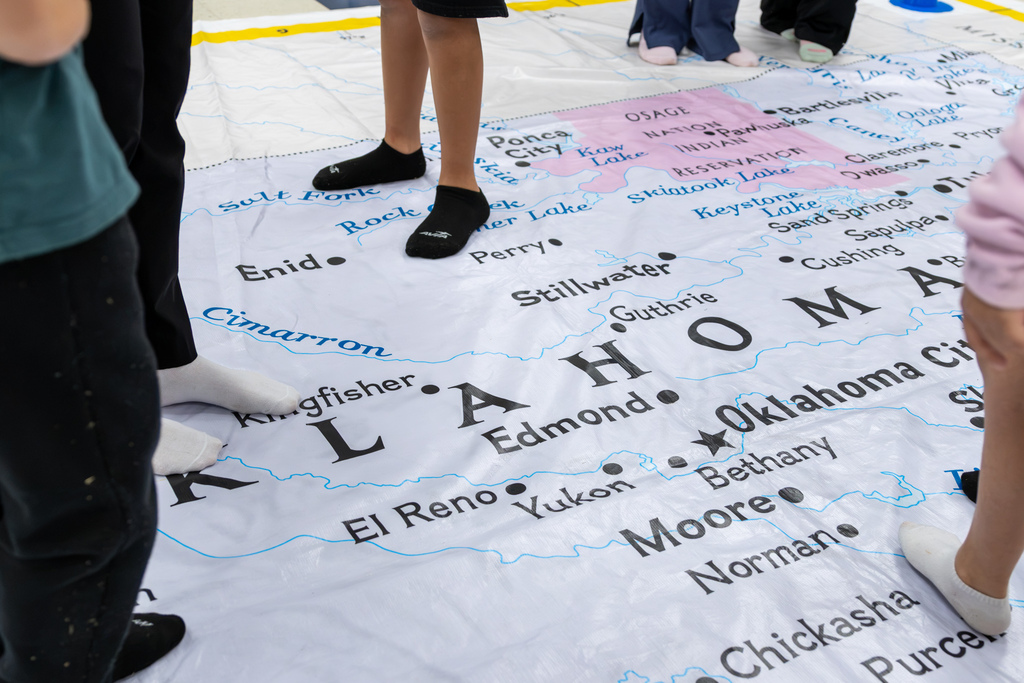 Students stand in their socks on a giant floor map of Oklahoma, with cities, rivers, lakes, and the Osage Nation reservation labeled across the map.