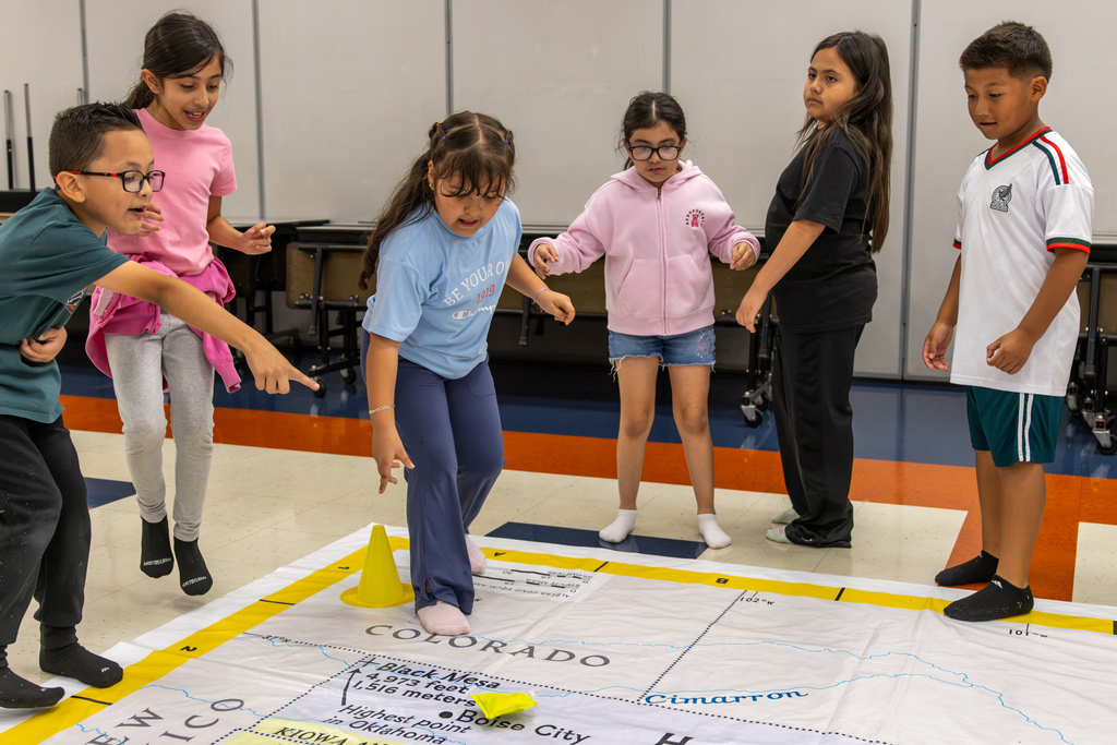 Students stand around the edge of the giant Oklahoma map and point toward locations near Colorado as they complete a geography activity.