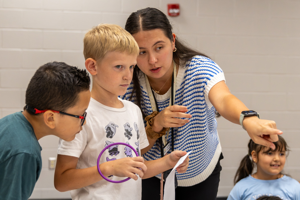 A teacher points toward the map while helping two students holding an activity card and a purple ring during the Oklahoma geography lesson.