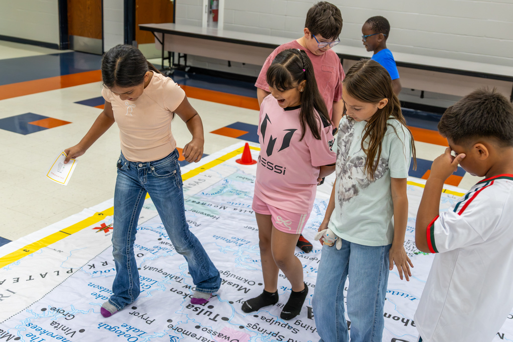 Central Elementary students gather on a giant Oklahoma map, looking down and stepping between labeled locations as they work through a geography activity.