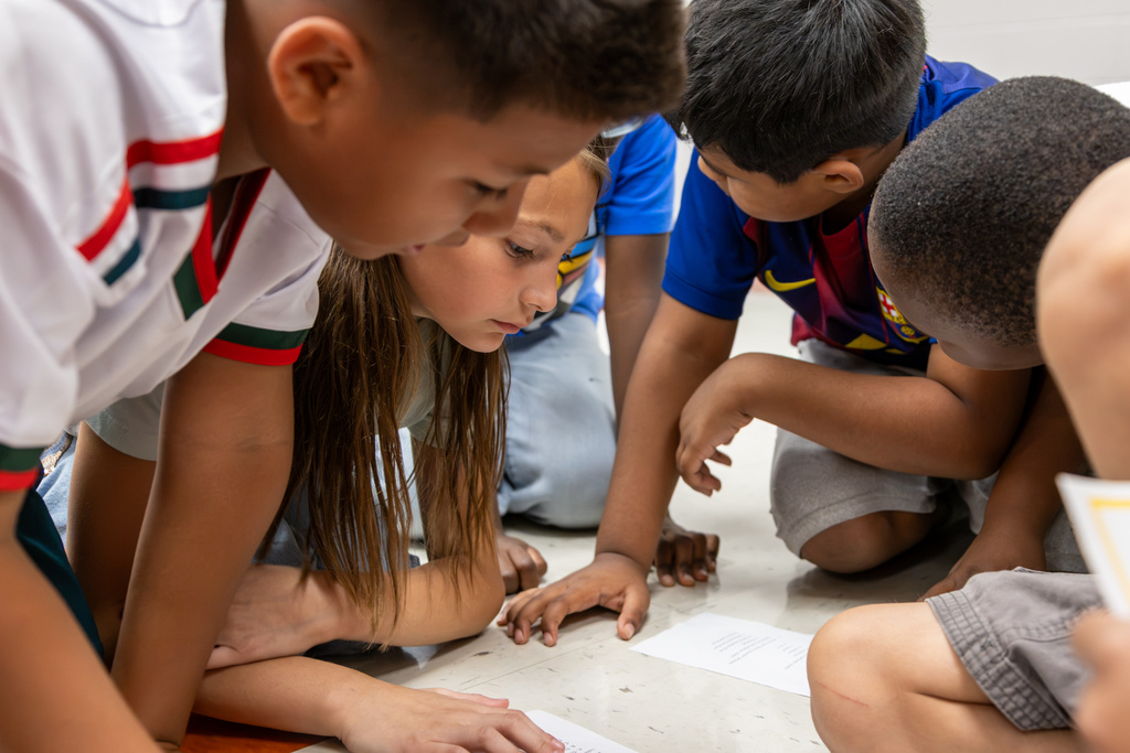 A group of students kneel closely together on the floor, reading activity cards during the giant Oklahoma map lesson.