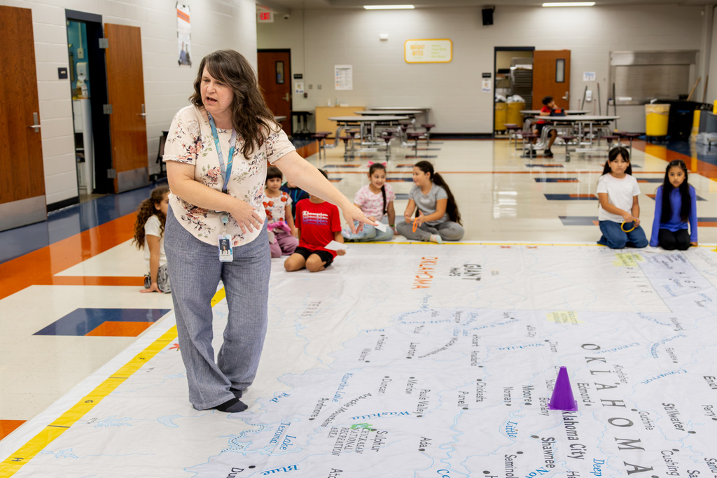 A teacher gestures across the giant Oklahoma map while students sit around its edges and listen to instructions.