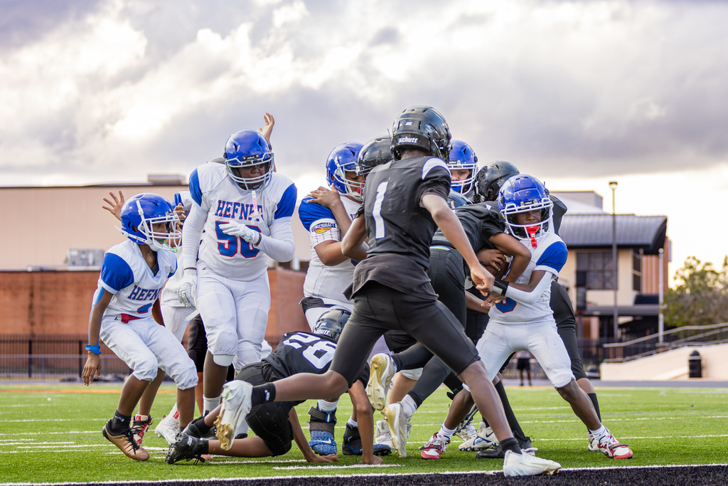Hefner and Cooper football players collide in a crowded goal-line play beneath a bright, cloudy sky as multiple players converge around the ball carrier.