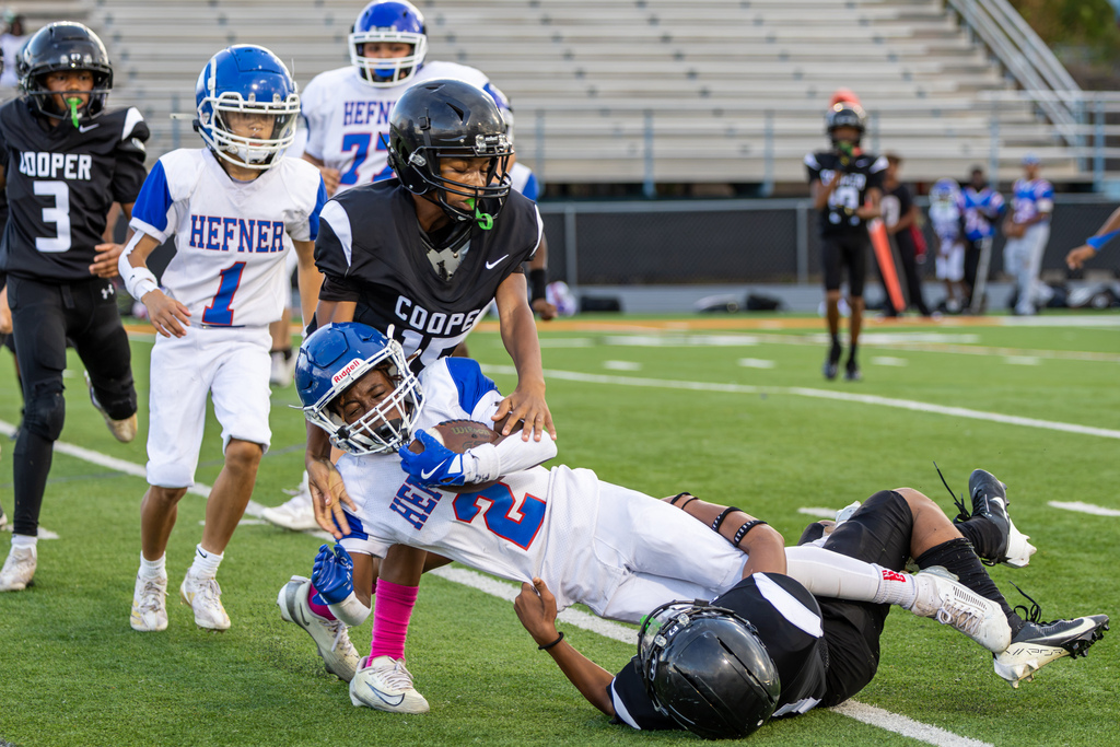 A Hefner Middle School player in white and blue is lifted sideways during a tackle by Cooper defenders while holding tightly to the football.