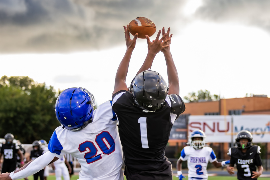 A Cooper Middle School football player in black and a Hefner Middle School player in white and blue reach high for the football during a contested catch under a dramatic cloudy sky.