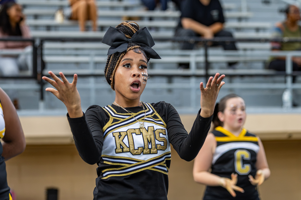 A Cooper Middle School cheerleader in a black, white, and gold uniform cheers with both hands raised and an expressive look on her face.
