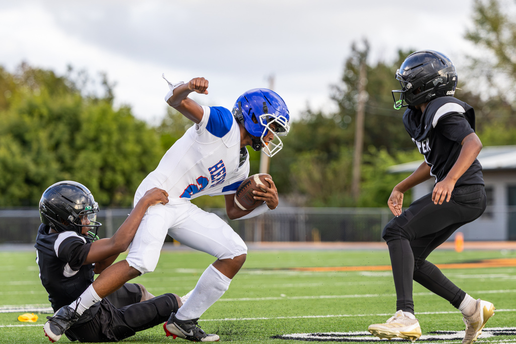 A Hefner Middle School football player in white and blue runs with the ball while a Cooper defender in black grabs at him from behind and other players pursue the play.