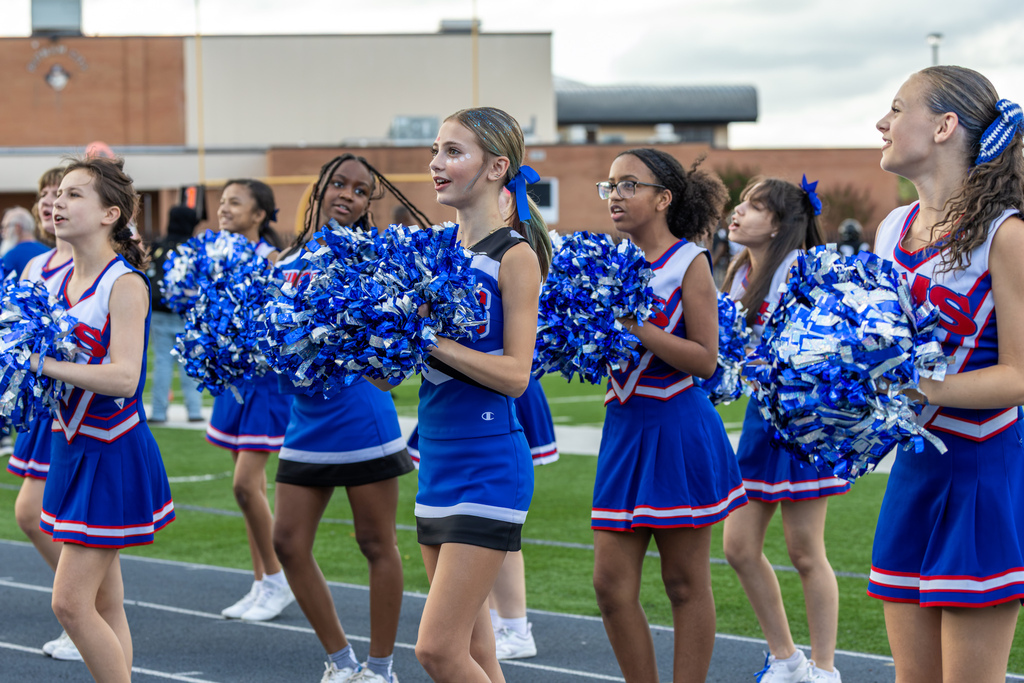 Hefner Middle School cheerleaders in blue, white, and red uniforms cheer from the sideline while holding blue and silver pom-poms.