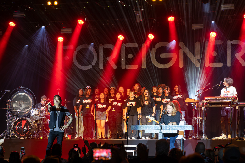 Putnam City High School choir students perform on stage behind members of Foreigner under bright red and white concert lights.