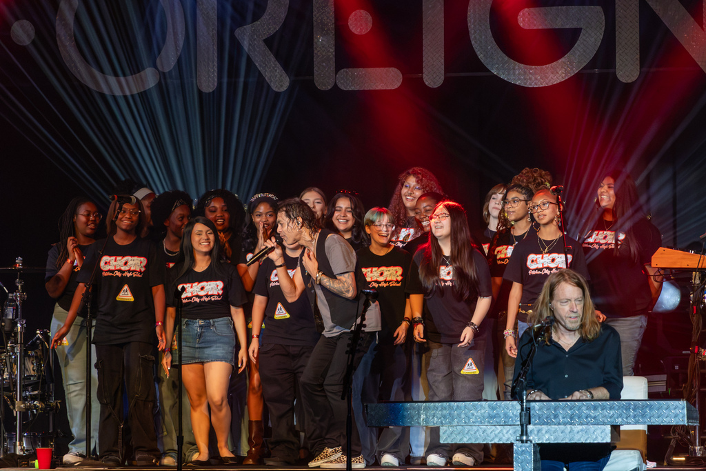 A Foreigner vocalist sings beside Putnam City High School choir students on stage while the students smile and sing along; a keyboard player performs in the foreground.