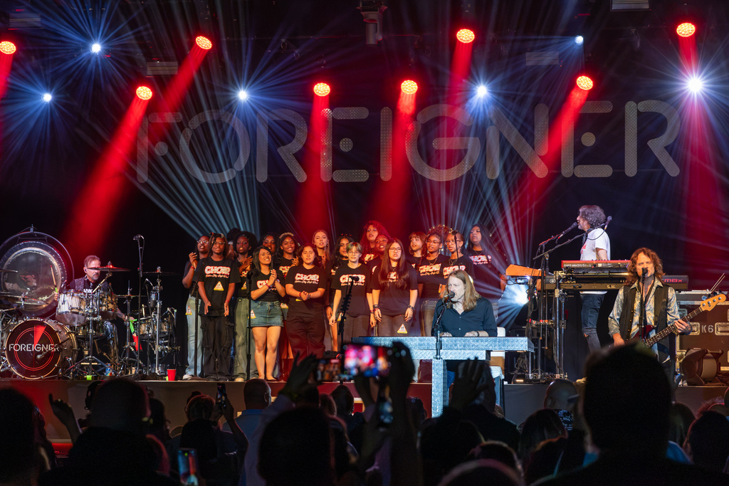 Putnam City High School choir sings with Foreigner on stage as red and white beams of light shine across the backdrop and audience members watch from below.