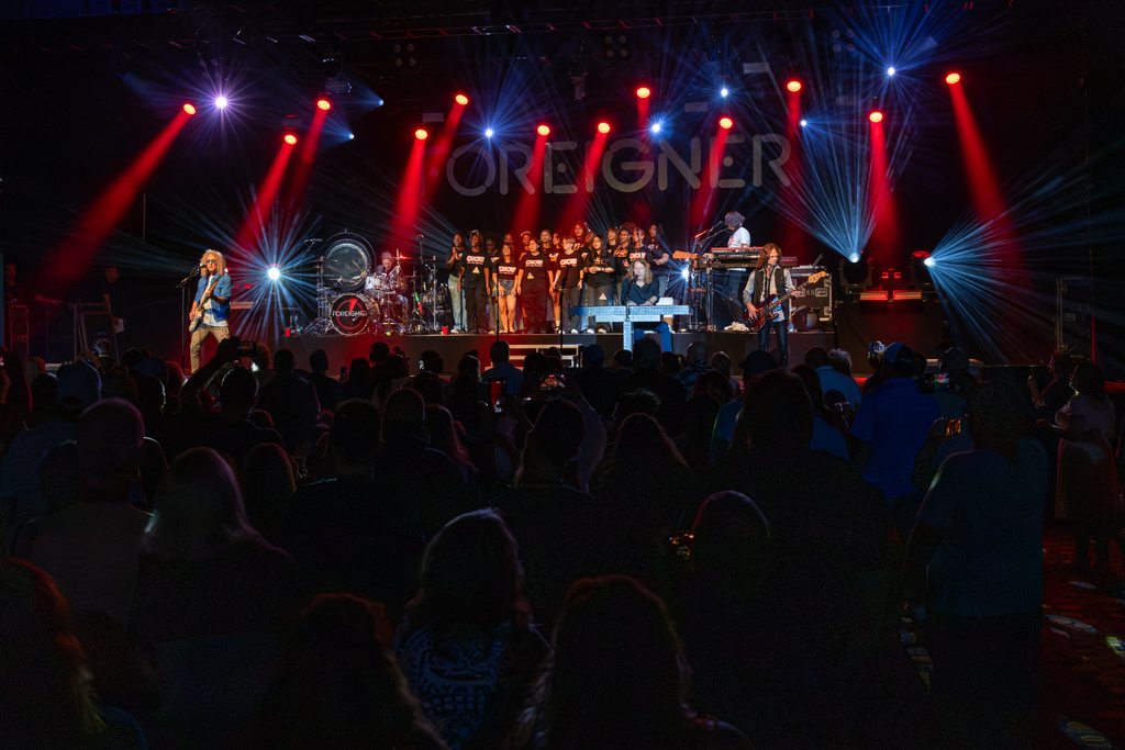 Wide view of the Foreigner concert stage with Putnam City High School choir performing at center, surrounded by band members, dramatic red and blue lighting, and a large audience.