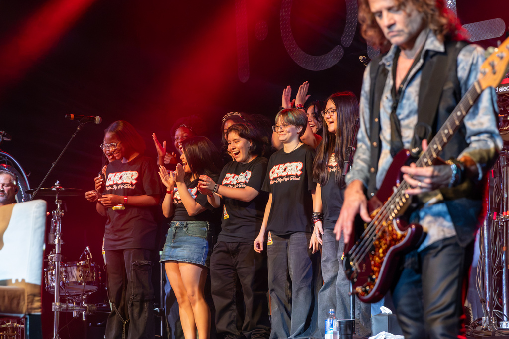 Putnam City High School choir students smile and clap on stage during the Foreigner concert while a guitarist performs in the foreground.