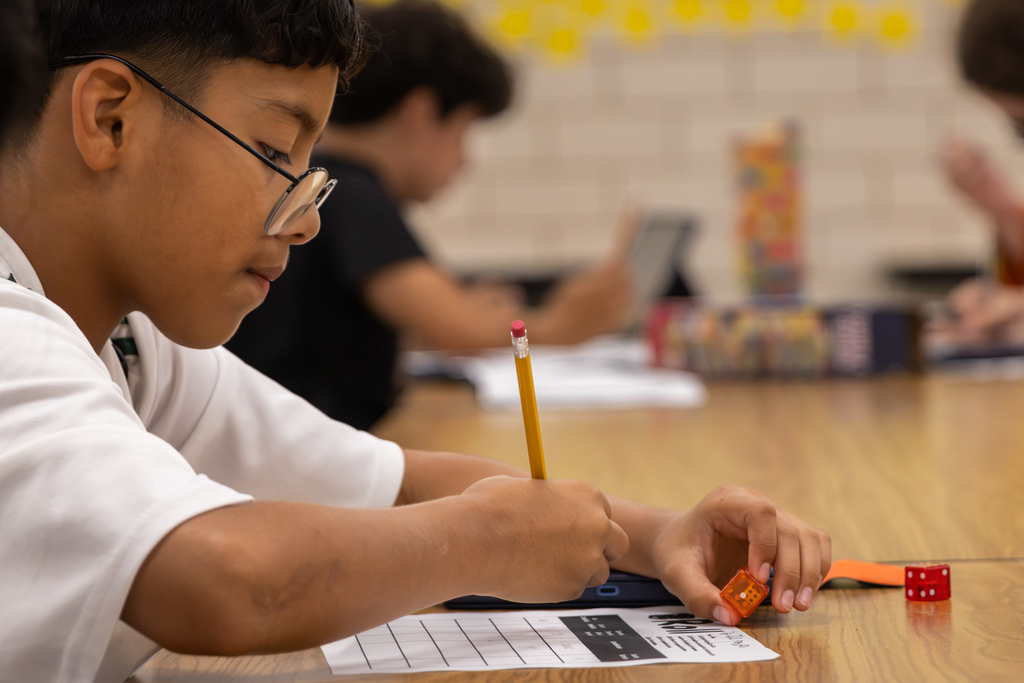 A Western Oaks Middle School student writes on a worksheet while holding a die during a classroom math game.