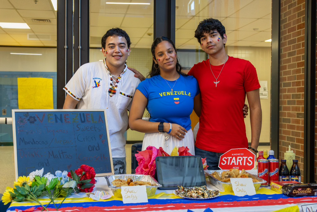 Three Putnam City West students stand behind a Venezuela-themed table with food samples, drinks, candy, flowers, and cultural displays.
