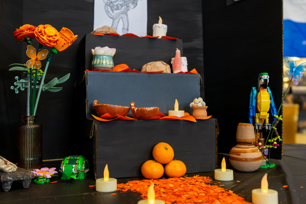 A Día de los Muertos display features candles, pottery, flowers, fruit, and decorative items arranged against a black backdrop.
