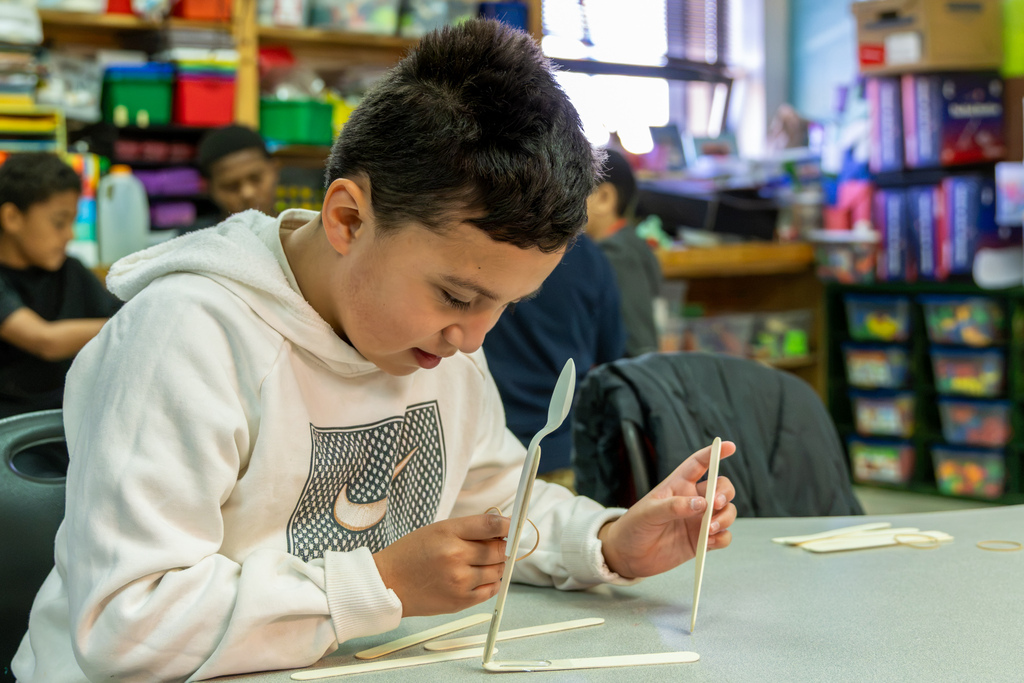 A Western Oaks Elementary student in a white hoodie concentrates on adjusting a rubber band attached to his handmade catapult.