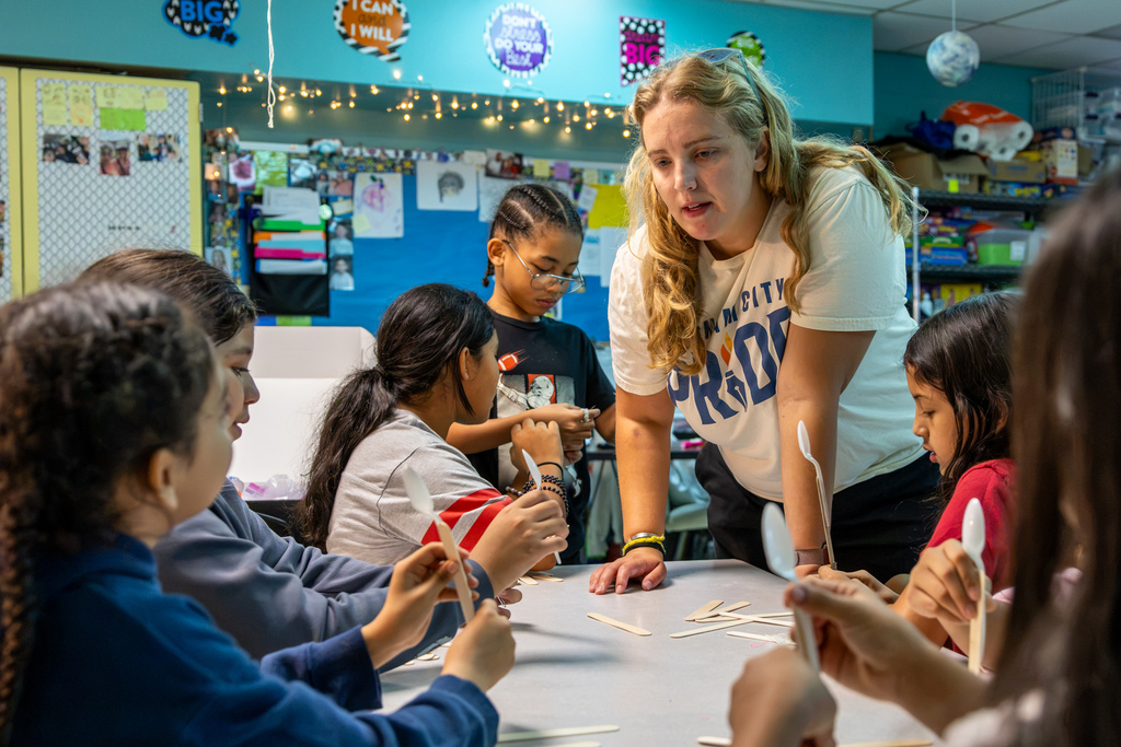 Mrs. Prahm leans over a classroom table to help Western Oaks Elementary students as they build catapults using plastic spoons and craft sticks.