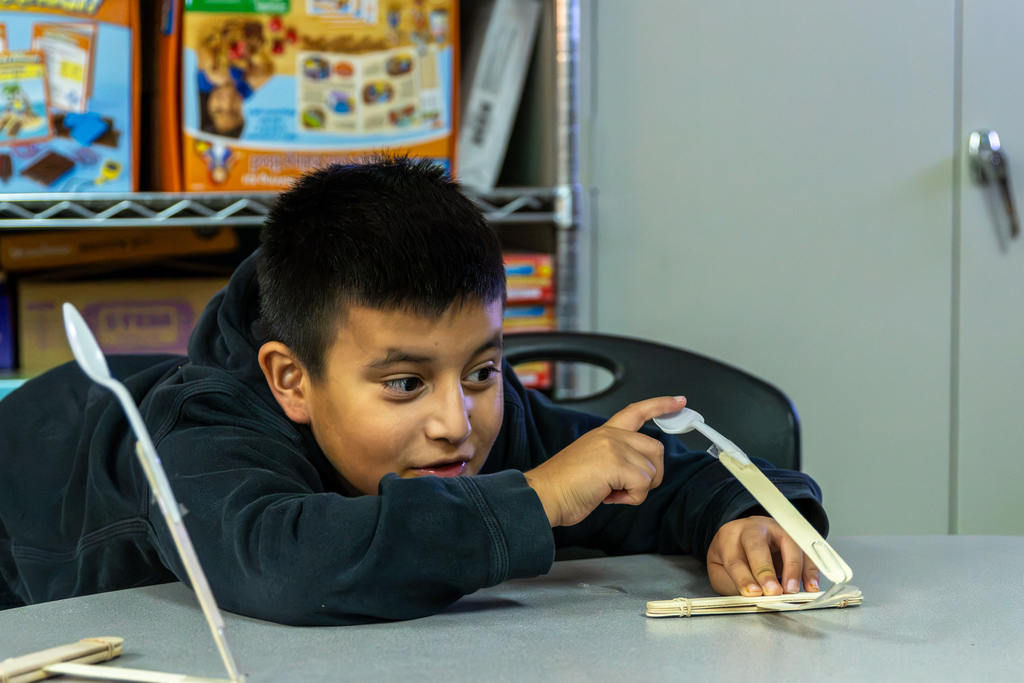 A Western Oaks Elementary student leans close to his catapult and points at its plastic spoon mechanism while examining his design.