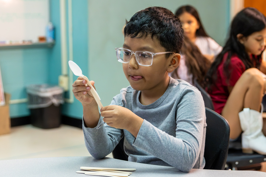 A Western Oaks Elementary student wearing glasses carefully attaches a plastic spoon to craft sticks while building a catapult in STEM class.