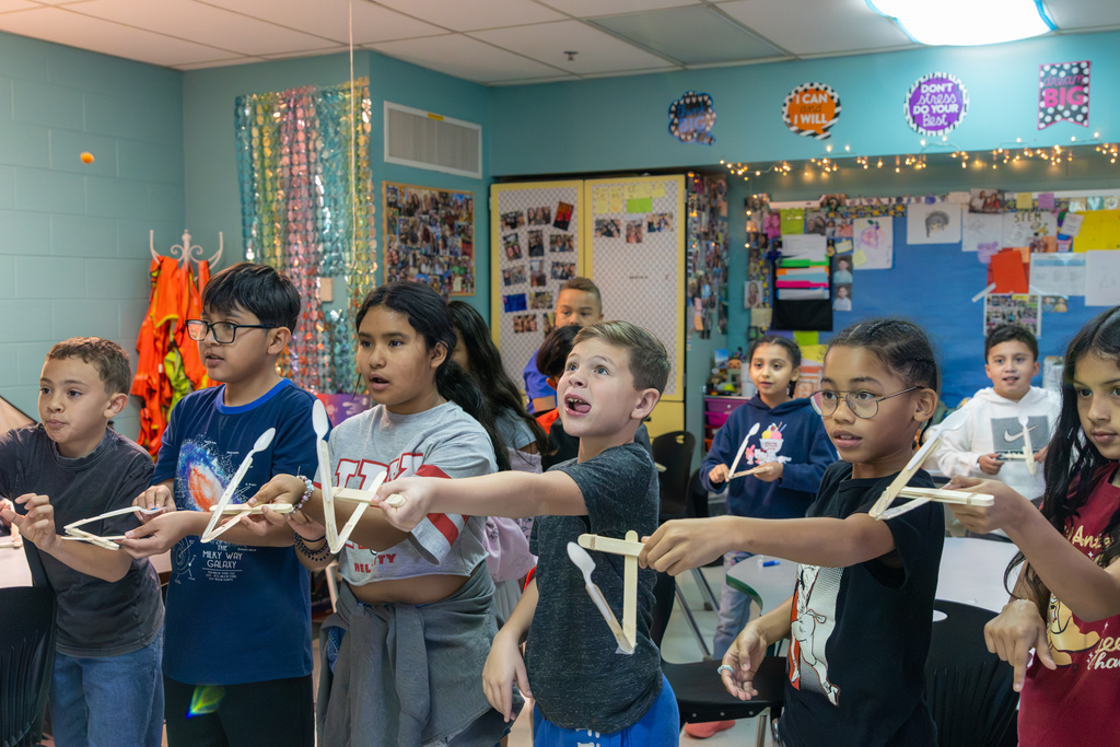 Western Oaks Elementary students stand together holding their handmade catapults, launching small objects across the STEM classroom.