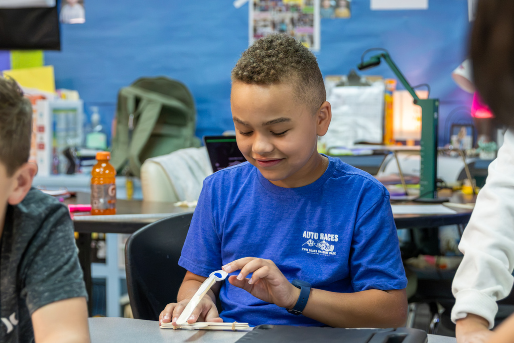 A Western Oaks Elementary student in a blue shirt smiles while adjusting the spoon mechanism on his craft-stick catapult.