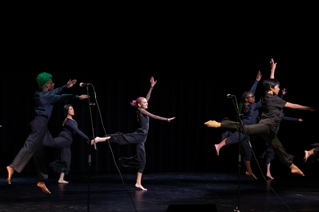 Student dancers leap and extend their arms across the stage during a contemporary dance performance, wearing black costumes against a dark backdrop.