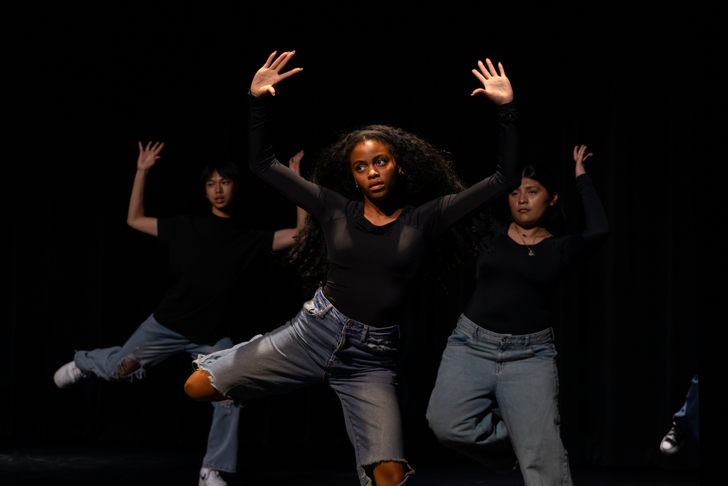 Three student dancers perform synchronized choreography with both arms raised, led by a student balancing on one leg at center stage.