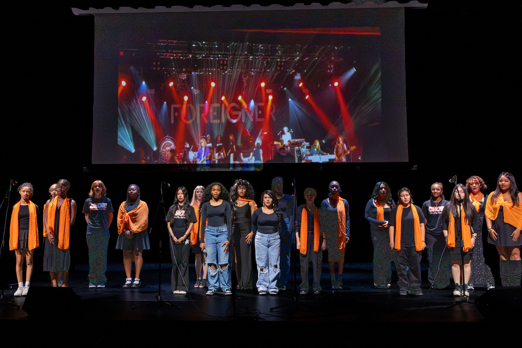 Putnam City High School choir students stand across the stage beneath a large projection of the band Foreigner performing under colorful concert lights.