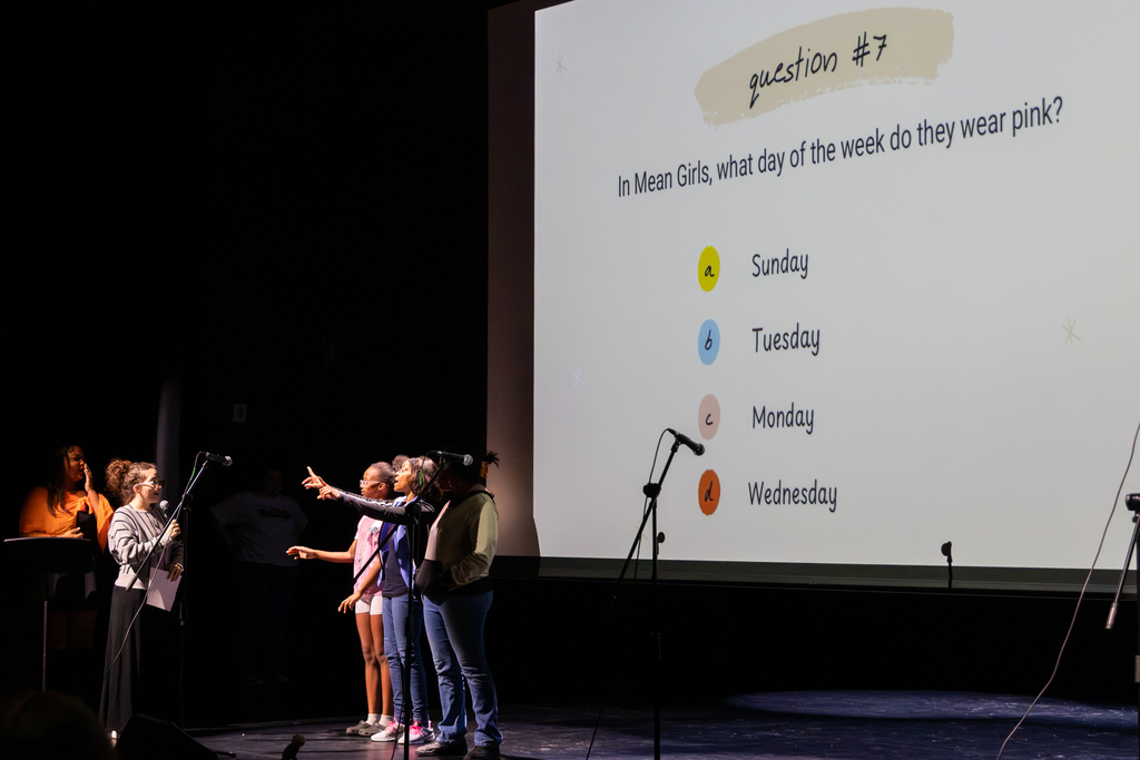 Students participate in a Mean Girls trivia activity onstage, pointing toward a projected multiple-choice question about which day the characters wear pink.