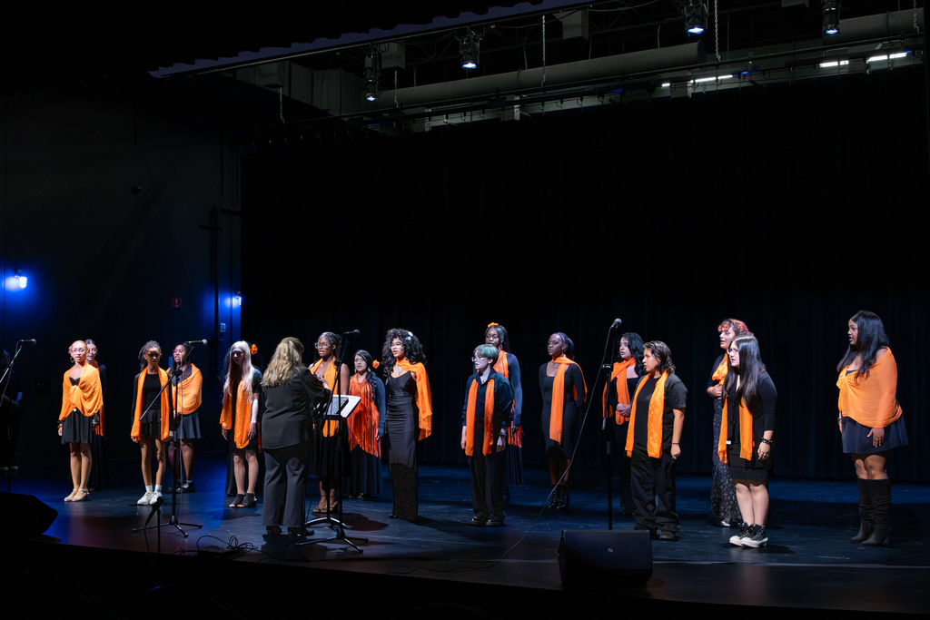 Putnam City High School choir members perform across the stage in black outfits accented with orange scarves as their director conducts from the front.