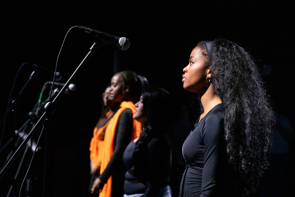 Side view of a student singer in black performing at a microphone, with fellow theatre members lined up behind her on a darkened stage.