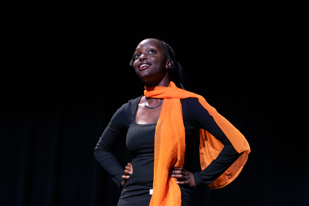 A smiling theatre student wearing a bright orange scarf stands confidently with her hands on her hips beneath a stage spotlight.
