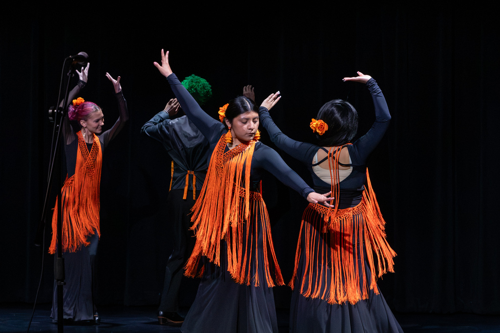 Four student dancers perform in flowing black costumes decorated with orange fringe, extending their arms in coordinated poses against a black stage curtain.