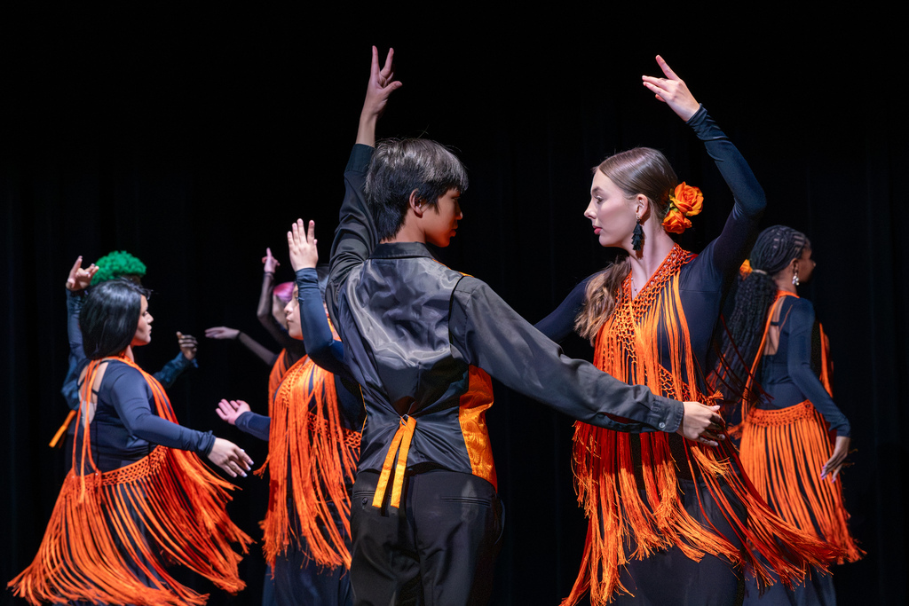 Student dancers in black costumes with bright orange fringe perform expressive choreography, facing one another with their arms raised.