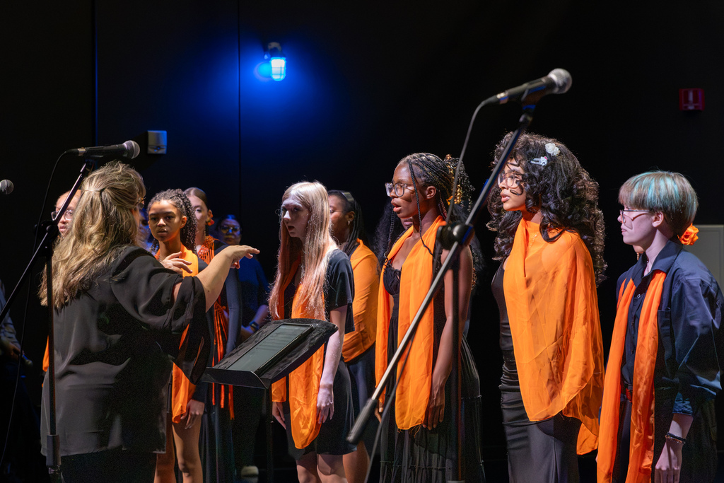 A choir director conducts Putnam City High School singers dressed in black and orange during a performance onstage.