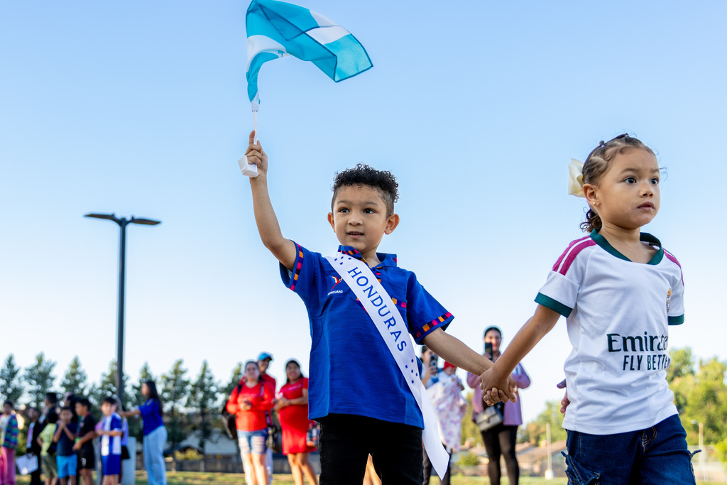 A young student wearing a blue Honduras soccer jersey and a sash reading “Honduras” waves a Honduran flag while holding hands with another student during the parade.