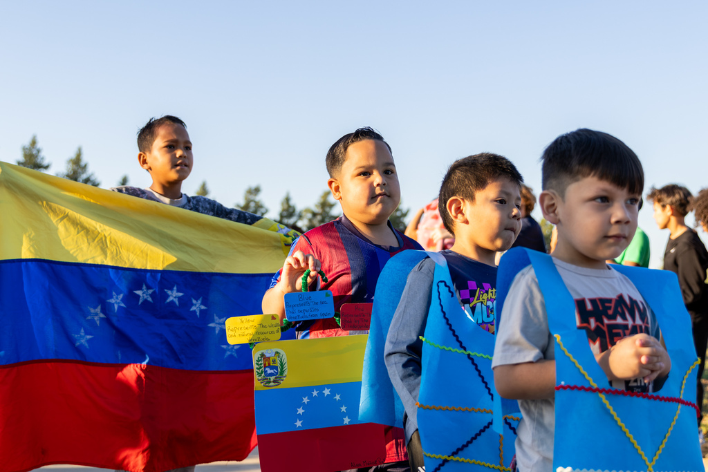 Elementary students walk in a parade carrying a large Venezuelan flag and wearing handmade blue costumes decorated with colorful ribbon.