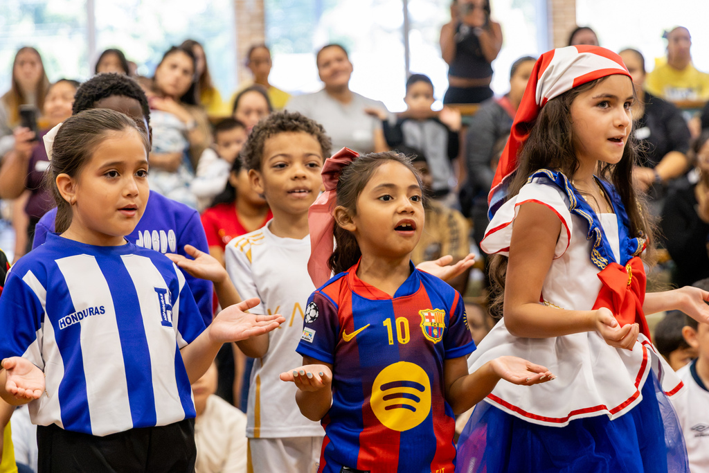 Three students wearing clothing representing Honduras and other cultural traditions sing and gesture with their hands during an indoor performance as families watch behind them.