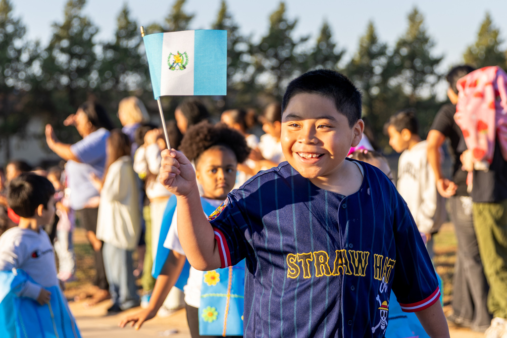 A smiling student in a navy blue baseball jersey proudly waves a small Guatemalan flag during the outdoor Hispanic Heritage Month parade.
