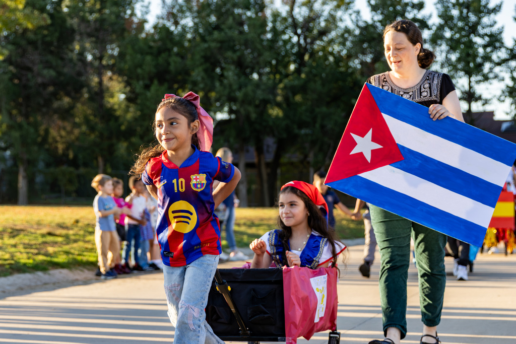 A smiling student wearing a soccer jersey walks at the front of the parade beside a younger student riding in a wagon, while an adult behind them carries a large Cuban flag.